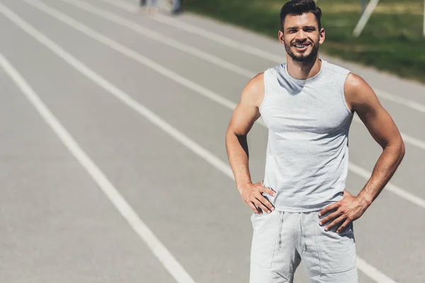 Sonriente joven deportista de pie en pista de atletismo con los brazos akimbo - foto de stock