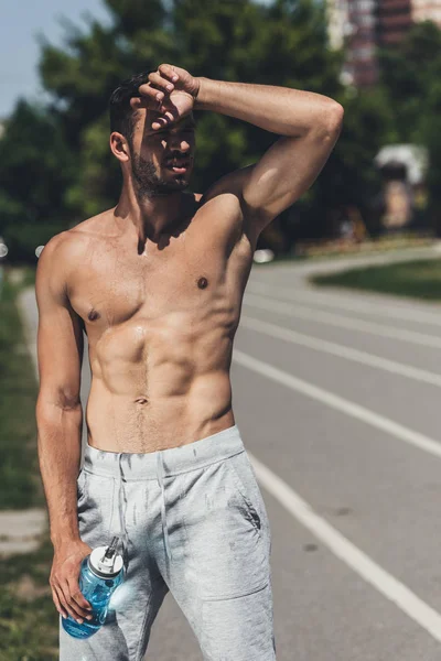 Молодой человек без рубашки, стоящий на беговой дорожке после тренировки и протирая лоб — стоковое фото