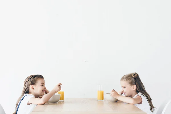 Adorable smiling schoolgirls eating cereals with orange juice for breakfast while sitting in front of each other isolated on white — Stock Photo