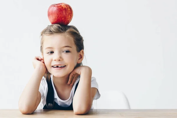 Pequena estudante sentada à mesa com maçã vermelha na cabeça isolada no branco — Fotografia de Stock