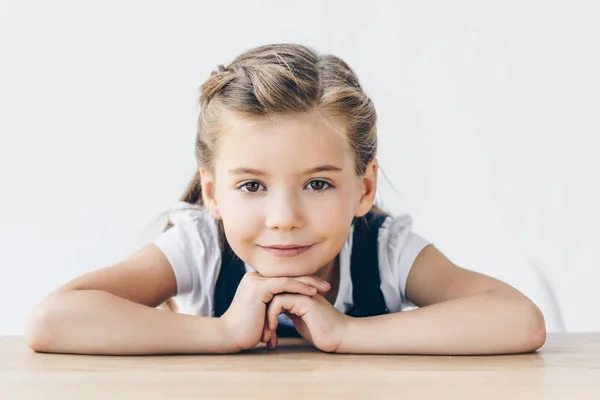 Улыбающаяся маленькая школьница, прислонившаяся к столу и смотрящая на камеру, изолированную на белом — стоковое фото
