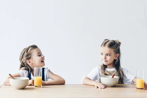 Очаровательные улыбающиеся школьницы едят хлопья с апельсиновым соком на завтрак изолированы на белом — стоковое фото