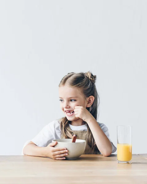 Pequena estudante feliz com tigela de café da manhã e suco de laranja isolado no branco — Fotografia de Stock