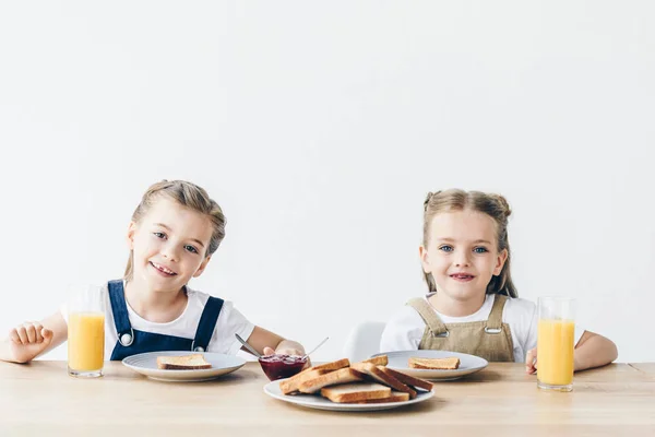 Hermanas pequeñas comiendo tostadas con mermelada para el desayuno y mirando a la cámara aislada en blanco - foto de stock