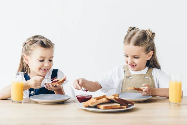 Усміхнені маленькі сестри їдять наносити варення на тости на сніданок ізольовані на білому — стокове фото