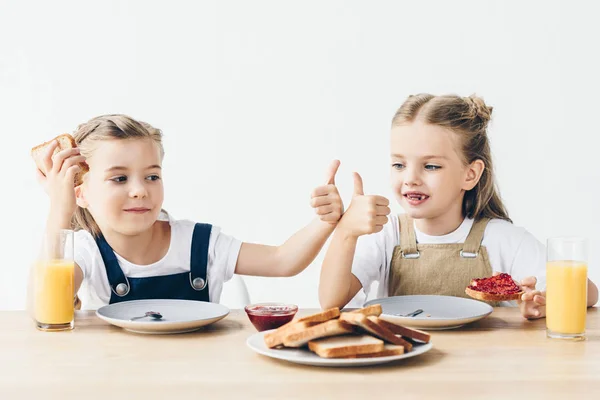 Petites sœurs manger des toasts avec confiture pour le petit déjeuner et montrant pouce vers le haut isolé sur blanc — Photo de stock