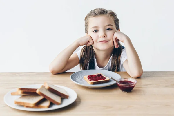 Sonriente colegiala con brindis y mermelada para el desayuno mirando a la cámara aislada en blanco - foto de stock