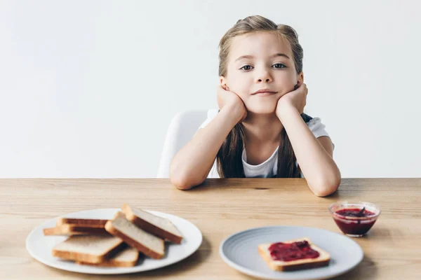 Задумлива маленька школярка з тостами і варенням для сніданку, дивлячись на камеру ізольовано на білому — стокове фото