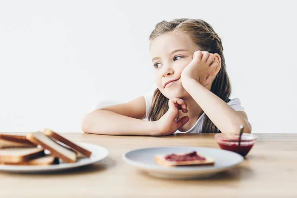 Nachdenkliches kleines Schulmädchen mit Toasts und Marmelade zum Frühstück, das isoliert auf weißem Grund wegschaut — Stockfoto