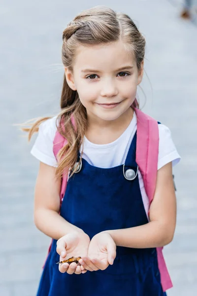 Entzückendes kleines Schulmädchen mit Schmetterling in den Händen und Blick in die Kamera — Stockfoto