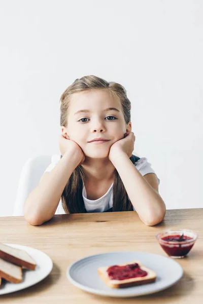 Little schoolgirl sitting at table and having toasts with jam for breakfast isolated on white — Stock Photo
