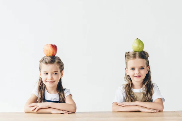 Adorable little schoolgirls with apple and pear on heads sitting at table isolated on white — Stock Photo