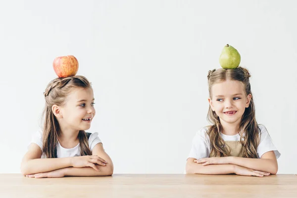 Colegialas felices con manzana y pera en las cabezas sentadas en la mesa aisladas en blanco - foto de stock