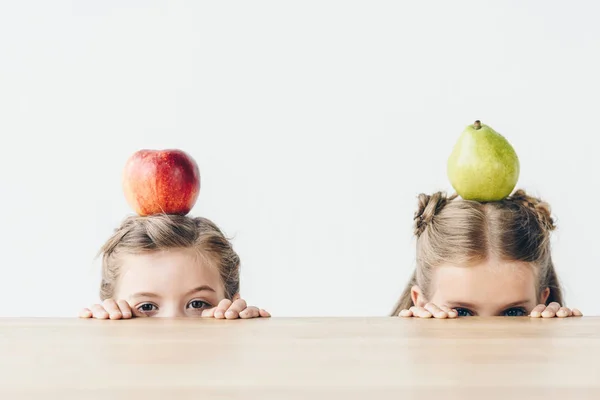 Colegiais com maçã e pêra na cabeça escondida atrás da mesa isolada no branco — Fotografia de Stock