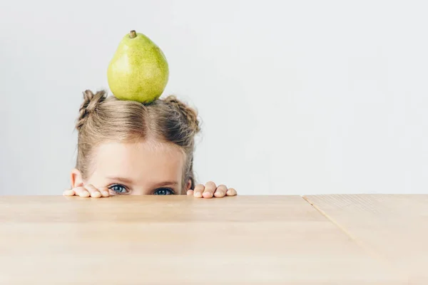 Little schoolgirl with ripe pear on head peeping from behind tabletop isolated on white — Stock Photo