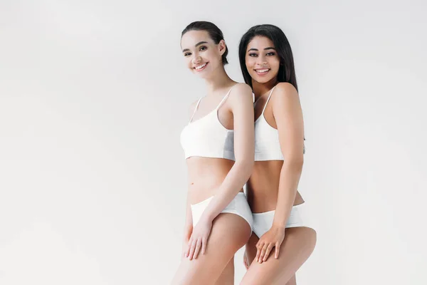 Donne multiculturali attraenti sorridenti in posa in lingerie bianca isolata su sfondo grigio — Foto stock