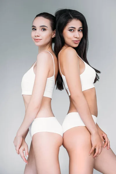 Multicultural women in white lingerie standing back to back isolated on gray background — Stock Photo