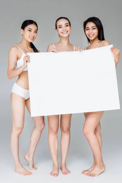 Femmes multiculturelles souriantes en lingerie tenant bannière vierge isolé sur fond gris — Photo de stock