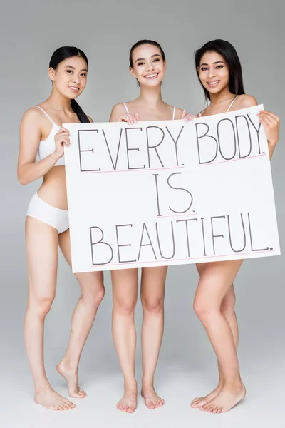 Souriant femmes multiculturelles tenant bannière avec lettrage chaque corps est belle isolé sur fond gris — Photo de stock