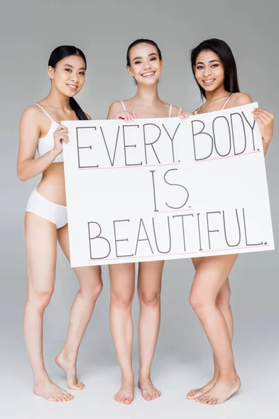 Mulheres multiculturais sorridentes segurando banner com letras cada corpo é bonito isolado em fundo cinza — Fotografia de Stock