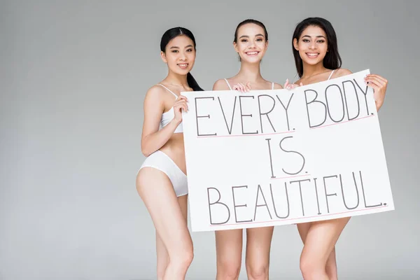 Sonrientes mujeres multiculturales sosteniendo pancarta con letras cada cuerpo es hermoso aislado sobre fondo gris - foto de stock