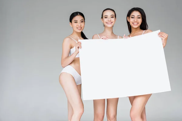 Tres sonrientes mujeres multiculturales en lencería sosteniendo banner en blanco aislado sobre fondo gris - foto de stock