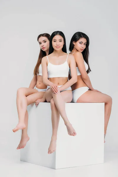 Muchachas multiétnicas en ropa interior sentadas en cubo blanco, aisladas en gris - foto de stock