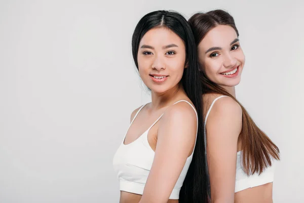 Attraktive lächelnde multiethnische Mädchen in weißen BHs, die in die Kamera schauen, isoliert auf grau — Stockfoto