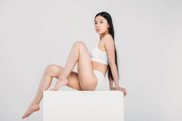 Элегантная азиатская девушка позирует в нижнем белье на белом кубе, изолированы на сером — стоковое фото