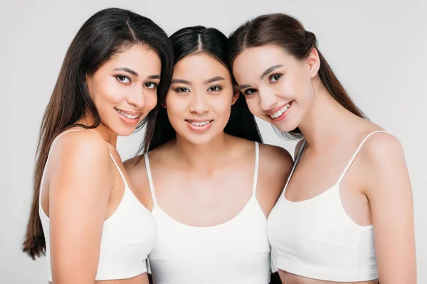 Щасливі мультикультурні дівчата позують у білих бюстгальтерах, ізольовані на сірому — стокове фото
