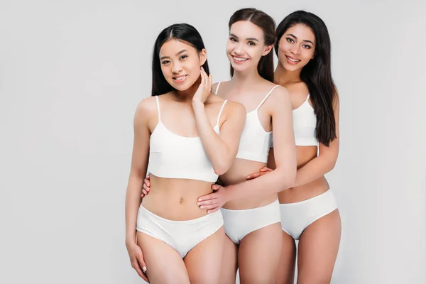 Souriant filles multiethniques étreignant et posant en lingerie blanche, isolé sur gris — Photo de stock