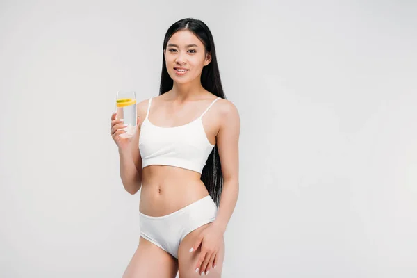 Attractive smiling asian girl holding glass of water, isolated on grey — Stock Photo