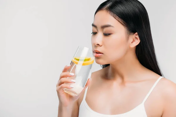 Encantadora chica asiática bebiendo agua pura con limón, aislado en gris - foto de stock