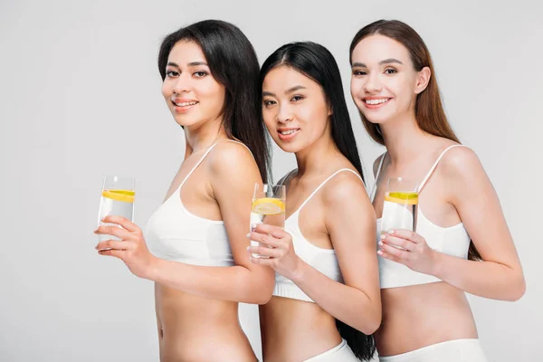 Hermosas chicas sonrientes multiétnicas sosteniendo vasos con agua de limón, aisladas en gris, estilo de vida saludable - foto de stock