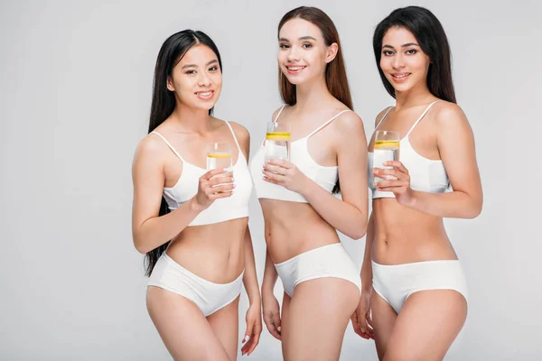 Elegant multicultural girls holding glasses with lemon water, isolated on grey, healthy lifestyle — Stock Photo