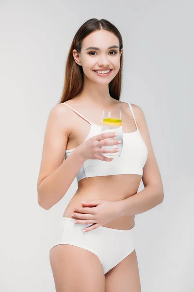 Симпатичная улыбающаяся девушка в белом нижнем белье, держащая стакан воды, изолированная на сером — стоковое фото