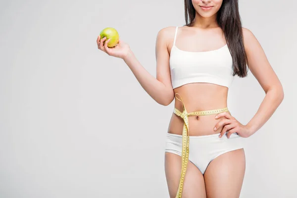 Обрезанный вид на молодую стройную женщину в нижнем белье с измерительной лентой на талии, держащую яблоко, изолированные на сером, диета концепции — стоковое фото