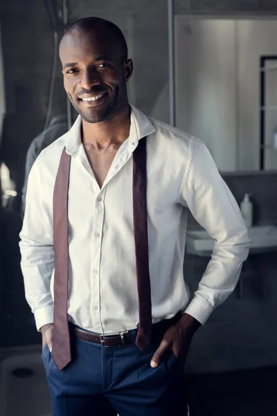 Smiling young businessman in white shirt with tie hanging on shoulders at bathroom — Stock Photo