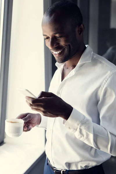 Sonriente joven hombre de negocios en camisa blanca usando teléfono inteligente durante el descanso de café - foto de stock
