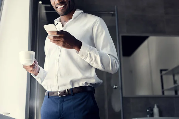 Recortado disparo de sonriente joven hombre de negocios en camisa blanca utilizando el teléfono inteligente durante el descanso de café - foto de stock