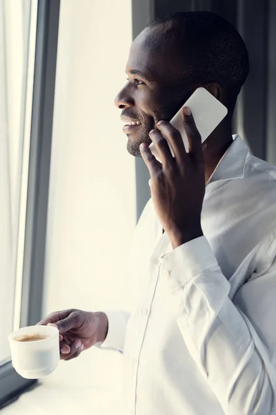 Happy young businessman in white shirt talking by phone during coffee break and looking through window — Stock Photo