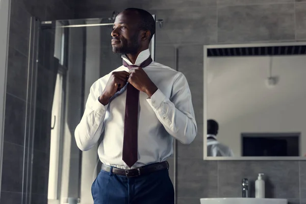 Joven hombre de negocios guapo en camisa blanca poniéndose la corbata en el baño - foto de stock