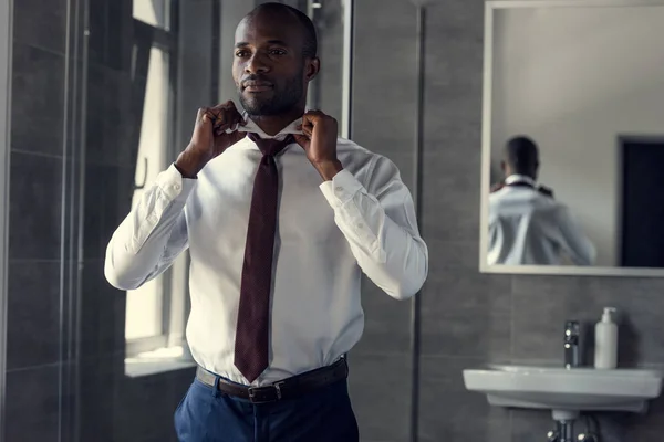 Счастливый молодой бизнесмен в белой рубашке надевает галстук в ванной комнате — стоковое фото