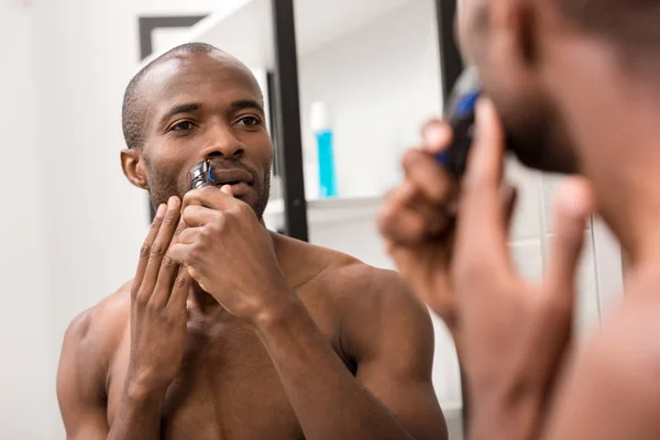 Atractivo joven afeitando la barba con afeitadora eléctrica mientras mira el espejo en el baño - foto de stock
