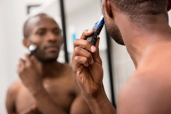 Recortado disparo de hombre joven afeitando la barba con afeitadora eléctrica mientras mira el espejo en el baño - foto de stock