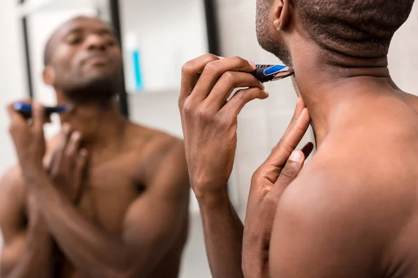 Africano americano joven afeitando la barba con afeitadora eléctrica mientras mira el espejo en el baño - foto de stock