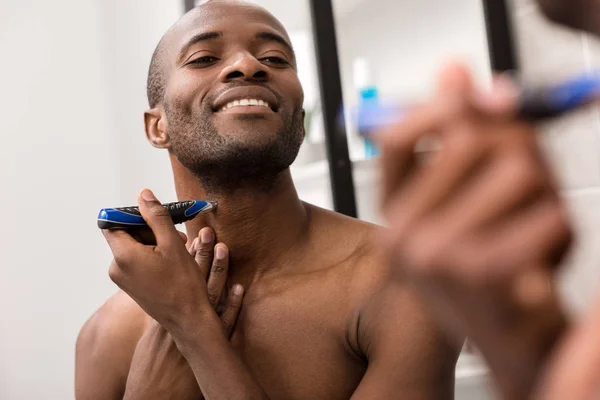 Sorridente jovem barbear barba com barbeador elétrico enquanto olha para o espelho no banheiro — Fotografia de Stock