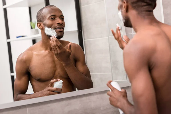 Привабливий молодий чоловік наносить гель для гоління, дивлячись на дзеркало у ванній — стокове фото