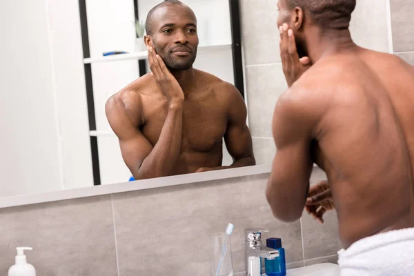 Beau jeune homme appliquant une lotion faciale après le rasage tout en regardant miroir dans la salle de bain — Photo de stock