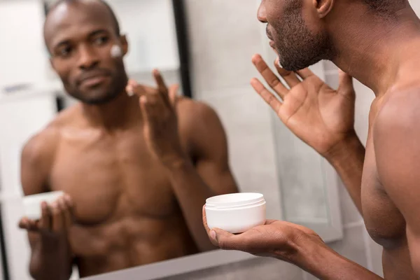 Beau jeune homme appliquant la crème faciale tout en regardant miroir dans la salle de bain — Photo de stock